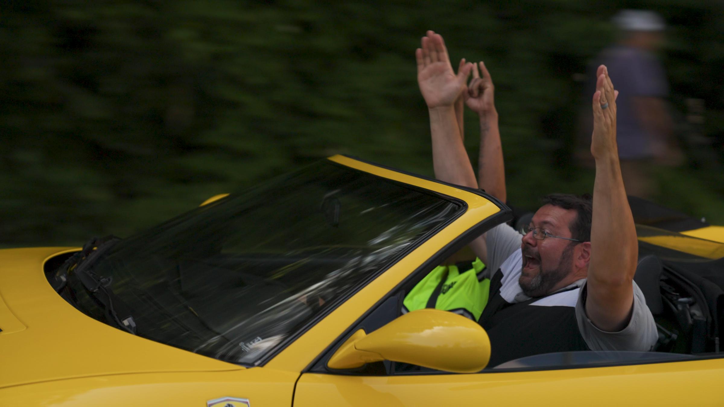Brad throws his arms up while riding in a yellow Ferrari